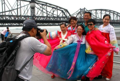 中国游客站在丹东的鸭绿江畔，穿上租来的朝鲜传统服，以鸭绿江断桥和对岸的朝鲜为背景，拍照留念。